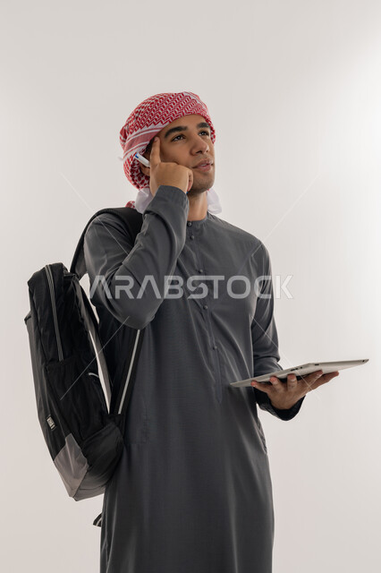 Planning and thinking about the future, finding solutions to problems and making decisions, using modern technical devices, an Arab Gulf Emirati university student wearing a kandura and turban, carrying a bag on his back, holding a tablet and placing his hand on his head with expressions of contemplation and concentration, gray background