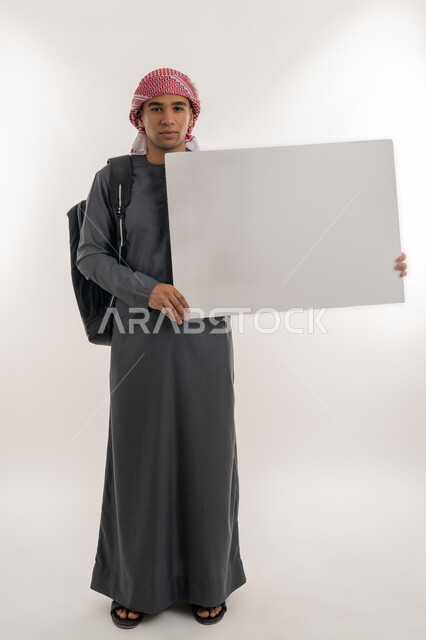 Announcing the start of a new university year in the Emirates, using explanatory panels and educational tools, empty rectangular mockup, portrait of an Arab Gulf Emirati university student wearing a kandura and turban holding a large white board in his hand, full-length body, gray background
