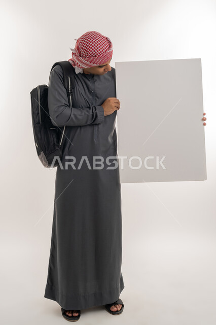 Using illustrations and educational tools, empty rectangle mockup, announcing the beginning of a new university year in the Emirates, portrait of an Arab Gulf Emirati university student wearing a kandura and turban holding a large white board in his hand, full-length body, gray background
