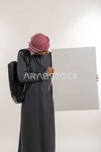 Using illustrations and educational tools, empty rectangle mockup, announcing the beginning of a new university year in the Emirates, portrait of an Arab Gulf Emirati university student wearing a kandura and turban holding a large white board in his hand, full-length body, gray background
