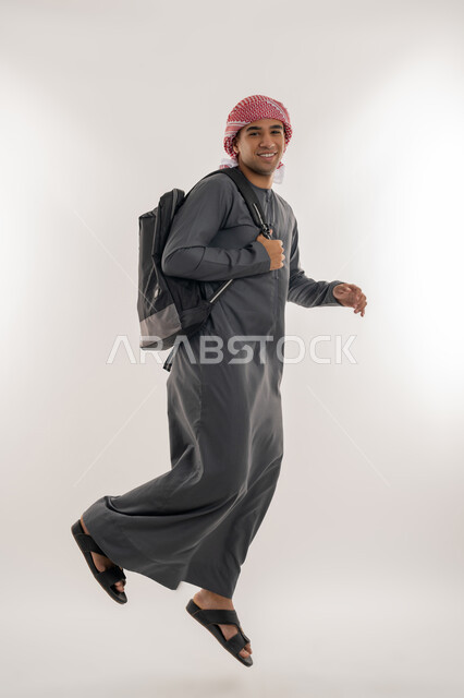 النظر إلى الكاميرا بإيماءات الفرح والسرور, الاستعداد للعام الجامعي الجديد, التحضير للذهاب إلى الجامعة, بورتريه لطالب جامعي عربي خليجي إماراتي يرتدي الكندورة والعمامة يحمل حقيبة على ظهره ويقفز عاليا بتعابير الحماس والسعادة, الجسم بالطول الكامل, خلفية رمادية