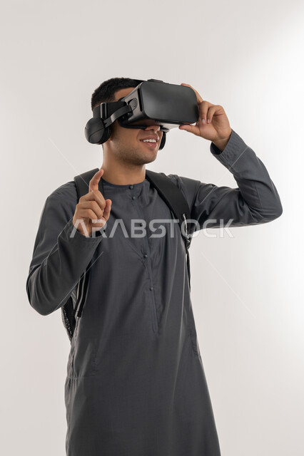 التعليم بواسطة خاصية الهولوجرام ثلاثية الابعاد, منصات تعليميه تعتمد على الواقع الافتراضي, محاكاة العالم الخيالي, بورتريه لطالب جامعي عربي خليجي إماراتي يرتدي الكندورة يضع نظارة الفي ار ويرفع اصبع السبابة لمحاولة لمس شيء ما, خلفية رمادية