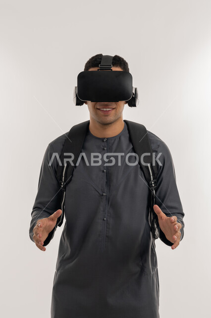 محاكاة العالم الخيالي, دمج الواقع الافتراضي بالتعلم والتدريب، استخدام التقنيات الحديثة في التعليم الأكاديمي, بورتريه لطالب جامعي عربي خليجي إماراتي يرتدي الكندورة يضع نظارة الفي ويرفع يديه بإيماءات التحجيم والإمساك بعنصر ما, خلفية رمادية