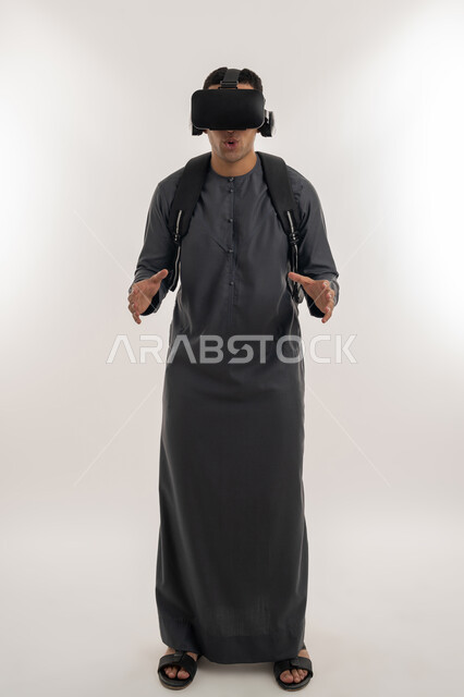 Using modern technologies in academic education, simulating the imaginary world, integrating virtual reality with learning and training, portrait of an Arab Gulf Emirati university student wearing a kandura, putting on glasses and raising his hands with gestures of scaling and holding an item, full-length body, gray background