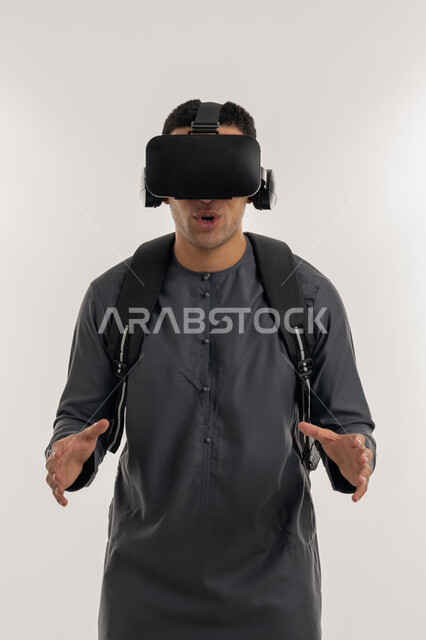 Advanced education quality using virtual world technology, simulating the imaginary world, integrating modern technologies with learning and training, portrait of an Arab Gulf Emirati university student wearing a kandura, wearing glasses and raising his hands with gestures of scaling and holding an item, gray background