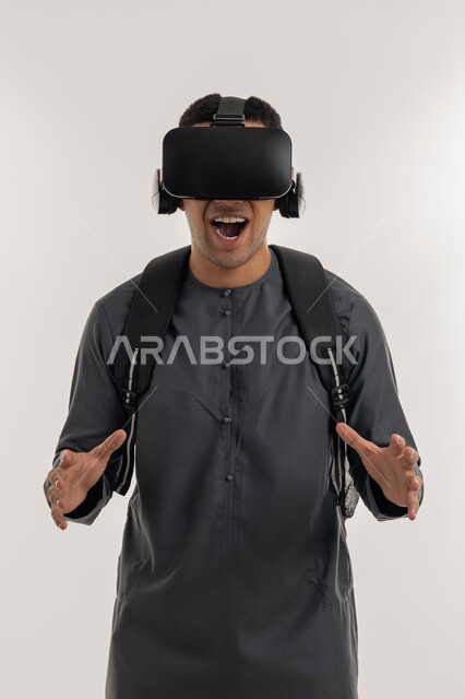 Advanced education quality using virtual world technology, simulating the imaginary world, integrating modern technologies with learning and training, portrait of an Arab Gulf Emirati university student wearing a kandura, wearing glasses and raising his hands with gestures of scaling and holding an item, gray background