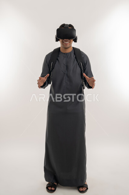 دمج الواقع الافتراضي بالتعلم والتدريب، محاكاة العالم الخيالي، استخدام التقنيات الحديثة في التعليم الأكاديمي, بورتريه لطالب جامعي عربي خليجي إماراتي يرتدي الكندورة يضع نظارة الفي ويرفع يديه بإيماءات التحجيم والإمساك بعنصر ما, الجسم بالطول الكامل، خلفية رمادية