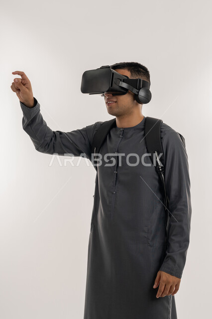 Wearing virtual reality glasses, using modern technologies in academic education, simulating the imaginary world, portrait of an Arab Gulf Emirati university student wearing a kandura, putting on 3D VR glasses and raising his hand to try to touch something, gray background