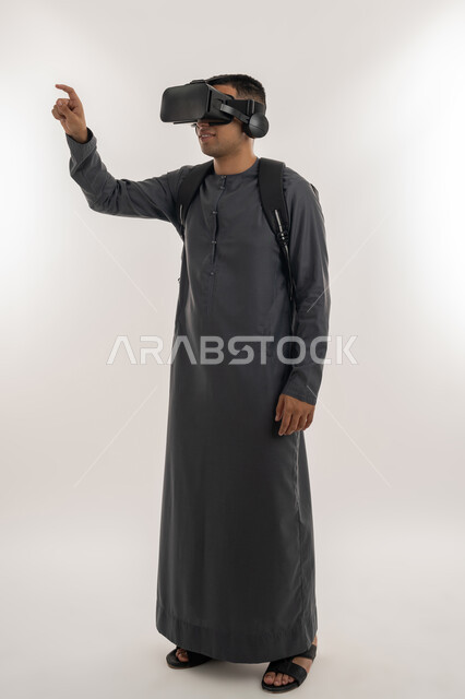 ارتداء نظارات الواقع الافتراضي, استخدام التقنيات الحديثة في التعليم الأكاديمي, محاكاة العالم الخيالي, بورتريه لطالب جامعي عربي خليجي إماراتي يرتدي الكندورة يضع نظارة الفي ار ثلاثية الأبعاد ويرفع يده لمحاولة لمس شيء ما, الجسم بالطول الكامل، خلفية رمادية