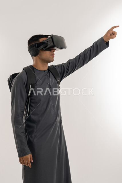 جودة التعليم المتطور باستخدام تقنية العالم الافتراضي, محاكاة العالم الخيالي، الإشارة بإصبع السبابة إلى شيء ما بالأعلى, بورتريه لطالب جامعي عربي خليجي إماراتي يرتدي الكندورة يحمل حقيبة على ظهره ويضع نظارة الفي ار ثلاثية الأبعاد, خلفية رمادية