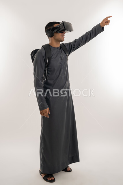 الإشارة بإصبع السبابة إلى شيء ما بالأعلى, دمج الواقع الافتراضي بالتعلم والتدريب، ارتداء نظارات العالم الخيالي, بورتريه لطالب جامعي عربي خليجي إماراتي يرتدي الكندورة يحمل حقيبة على ظهره ويضع نظارة الفي ار ثلاثية الأبعاد, الجسم بالطول الكامل، خلفية رمادية
