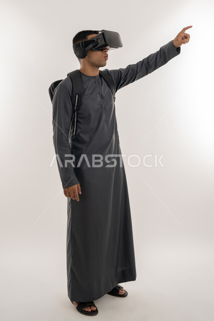 الإشارة بإصبع السبابة إلى شيء ما بالأعلى, دمج الواقع الافتراضي بالتعلم والتدريب، ارتداء نظارات العالم الخيالي, بورتريه لطالب جامعي عربي خليجي إماراتي يرتدي الكندورة يحمل حقيبة على ظهره ويضع نظارة الفي ار ثلاثية الأبعاد, الجسم بالطول الكامل، خلفية رمادية