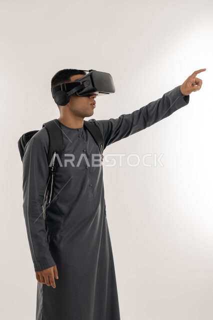 دمج الواقع الافتراضي بالتعلم والتدريب, الإشارة بإصبع السبابة إلى شيء ما بالأعلى، ارتداء نظارات العالم الخيالي, بورتريه لطالب جامعي عربي خليجي إماراتي يرتدي الكندورة يحمل حقيبة على ظهره ويضع نظارة الفي ار ثلاثية الأبعاد, خلفية رمادية