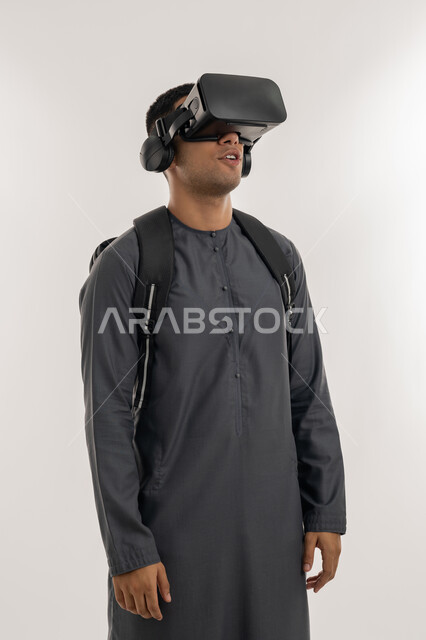 جودة التعليم المتطور باستخدام تقنية العالم الافتراضي, محاكاة العالم الخيالي، دمج التقنيات الحديثة بالتعلم والتدريب, بورتريه لطالب جامعي عربي خليجي إماراتي يرتدي الكندورة يحمل حقيبة على ظهره ويضع نظارة الفي ار ثلاثية الأبعاد, خلفية رمادية