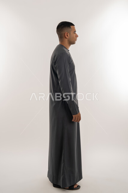 الاهتمام بالأناقة والمظهر الخارجي, مفهوم الرجولة والثقة بالنفس, بورتريه من الجانب لشاب عربي خليجي إماراتي يرتدي الكندورة يقف باستقامة واعتدال, الجسم بالطول الكامل, خلفية رمادية