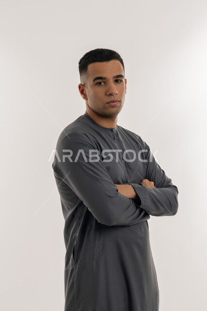 النظر الكاميرا بإيماءات الجدية والحزم, الاهتمام بالأناقة والمظهر الخارجي, تعابير الثقة والاعتزاز بالنفس, بورتريه لشاب عربي خليجي إماراتي يرتدي الكندورة يقف باستقامة واعتدال وأيدي متقاطعة, خلفية رمادية