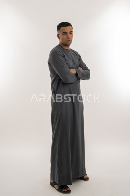 النظر الكاميرا بإيماءات الجدية والحزم, الاهتمام بالأناقة والمظهر الخارجي, تعابير الثقة والاعتزاز بالنفس, بورتريه لشاب عربي خليجي إماراتي يرتدي الكندورة يقف باستقامة واعتدال وأيدي متقاطعة, الجسم بالطول الكامل, خلفية رمادية