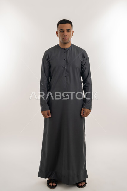 Looking at the camera with gestures of happiness and pleasure, interest in elegance and appearance, expressions of confidence and self-esteem, portrait of a young Arab Gulf Emirati man wearing a kandura standing straight and upright, full-length body, gray background