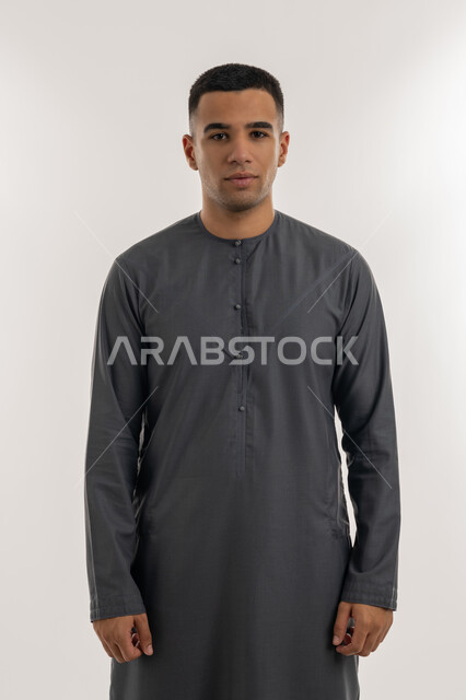 النظر إلى الكاميرا بإيماءات السعادة والسرور, الاهتمام بالأناقة والمظهر الخارجي, تعابير الثقة و الاعتزاز بالنفس, بورتريه لشاب عربي خليجي إماراتي يرتدي الكندورة  يقف باستقامة واعتدال, خلفية رمادية