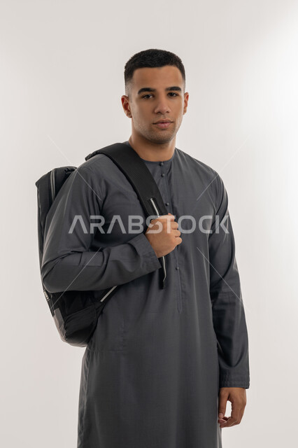 Caring about elegance and appearance, preparing for the new academic year, preparing to go to university, academic universities and colleges in the Emirates, portrait of an Arab Gulf Emirati university student wearing a kandura standing straight and carrying a bag on his shoulder, gray background