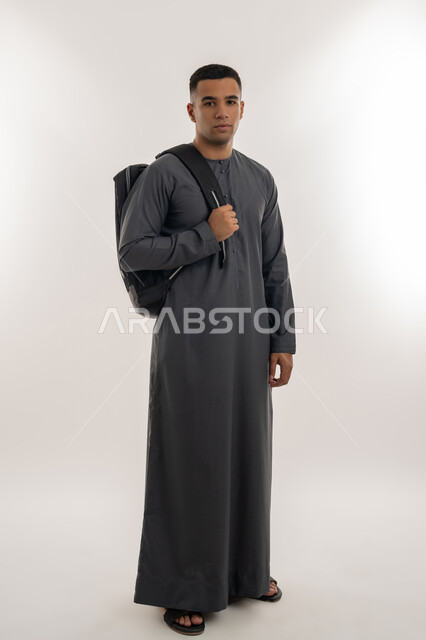 الاستعداد للعام الجامعي الجديد, التحضير للذهاب إلى الجامعة, الجامعات والكليات الأكاديمية في الإمارات, الاهتمام بالأناقة والمظهر الخارجي, بورتريه لطالب جامعي عربي خليجي إماراتي يرتدي الكندورة يقف باستقامة ويحمل حقيبة على كتفه, الجسم بالطول الكامل, خلفية رمادية