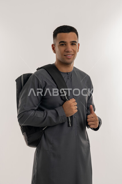النظر إلى الكاميرا بإيماءات السعادة والسرور, تعابير الجودة و الامتياز, التعليم الجامعي في الإمارات العربية المتحدة, بورتريه لطالب جامعي عربي خليجي إماراتي يرتدي الكندورة يحمل حقيبة على ظهره ويشير بإصبع الإبهام للأعلى, خلفية رمادية