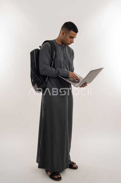 دمج التقنيات الحديثة بالتعليم الأكاديمي, المناهج والمواد التعليمية, التعليم العالي والجامعات في الإمارات, بورتريه لطالب جامعي عربي خليجي إماراتي يرتدي الكندورة يحمل حقيبة على ظهره ويقوم باستعمال جهاز الحاسوب المحمول, الجسم بالطول الكامل, خلفية رمادية