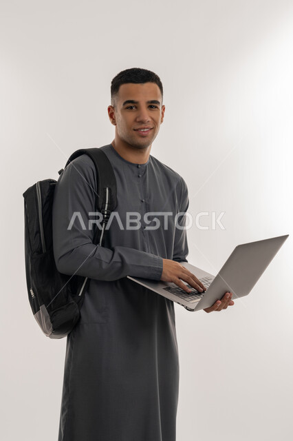 المناهج والمواد التعليمية, دمج التقنيات الحديثة بالتعليم الأكاديمي, التعليم العالي والجامعات في الإمارات, بورتريه لطالب جامعي عربي خليجي إماراتي يرتدي الكندورة يحمل حقيبة على ظهره ويقوم باستعمال جهاز الحاسوب المحمول, خلفية رمادية