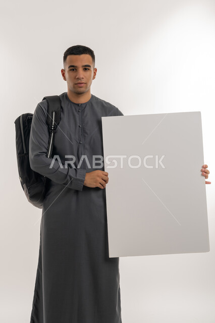 التعليم العالي في الجامعات والكليات الأكاديمية الإماراتية, استخدام لوحات توضيحية ووسائل تعليمية, موك اب مستطيل فارغ، بورتريه لطالب جامعي عربي خليجي إماراتي يرتدي الكندورة يحمل حقيبة على ظهره وبيده لوح أبيض كبير, خلفية رمادية