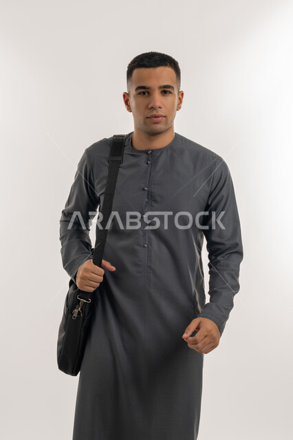 التحضير للذهاب إلى الجامعة, الجامعات والكليات الأكاديمية في الإمارات, الاهتمام بالمظهر الخارجي, الاستعداد للعام الجامعي الجديد, بورتريه لطالب جامعي عربي خليجي إماراتي يرتدي الكندورة ينظر إلى الكاميرا ويحمل حقيبة على كتفه, خلفية رمادية