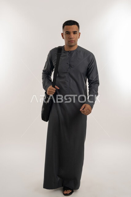 Preparing to go to university, academic universities and colleges in the Emirates, taking care of the external appearance, preparing for the new university year, portrait of an Arab Gulf Emirati university student wearing a kandura looking at the camera and carrying a bag on his shoulder, full body, gray background