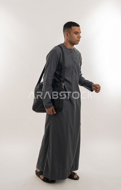 Attention to elegance and appearance, preparing for the new academic year, preparing to go to university, academic universities and colleges in the Emirates, portrait of an Arab Gulf Emirati university student wearing a kandura standing straight and carrying a bag on his shoulder, full-length body, gray background