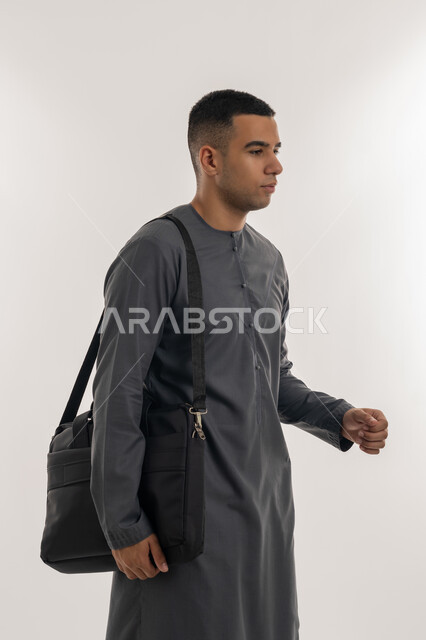 الاهتمام بالأناقة والمظهر الخارجي, الاستعداد للعام الجامعي الجديد, التحضير للذهاب إلى الجامعة, الجامعات والكليات الأكاديمية في الإمارات, بورتريه لطالب جامعي عربي خليجي إماراتي يرتدي الكندورة  يقف باستقامة ويحمل حقيبة على كتفه, خلفية رمادية