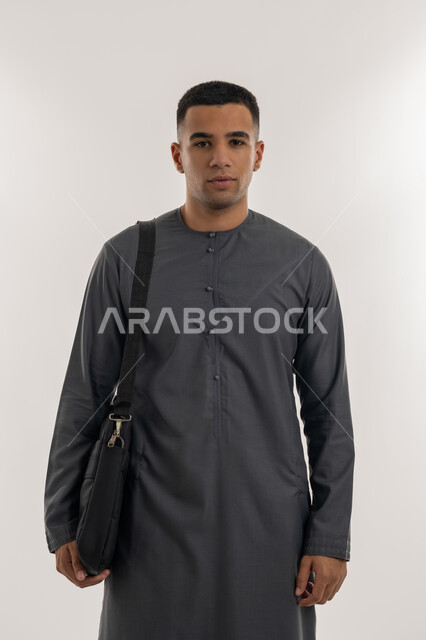الاهتمام بالأناقة والمظهر الخارجي, الاستعداد للعام الجامعي الجديد, التحضير للذهاب إلى الجامعة, الجامعات والكليات الأكاديمية في الإمارات, بورتريه لطالب جامعي عربي خليجي إماراتي يرتدي الكندورة  يقف باستقامة ويحمل حقيبة على كتفه, خلفية رمادية