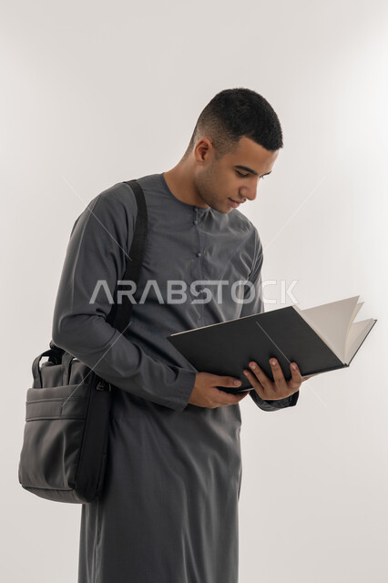 مراجعة الدروس اليومية, مفهوم الاجتهاد والتفوق, التعليم الجامعي في الإمارات العربية المتحدة, بورتريه لطالب جامعي عربي خليجي إماراتي يرتدي الكندورة يحمل بيده كتاب ويقوم بدراسة المحاضرات, خلفية رمادية