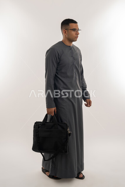 الاستعداد للعام الجامعي الجديد, التحضير للذهاب إلى الجامعة, التعليم العالي في الجامعات والكليات الأكاديمية الإماراتية, بورتريه لطالب عربي خليجي إماراتي يرتدي الكندورة يضع نظارة طبية ويحمل بيده حقيبة, الجسم بالطول الكامل, خلفية رمادية