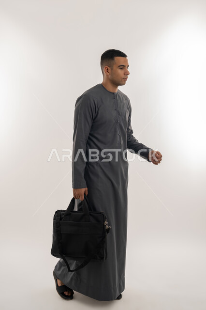 التحضير للذهاب إلى الجامعة, الاستعداد للعام الجامعي الجديد, التعليم العالي في الجامعات والكليات الأكاديمية الإماراتية, بورتريه لطالب عربي خليجي إماراتي يرتدي الكندورة يضع نظارة طبية ويحمل بيده حقيبة, الجسم بالطول الكامل, خلفية رمادية