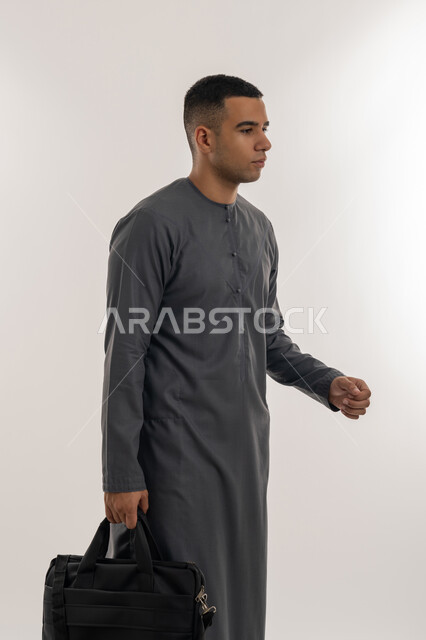 التعليم العالي في الجامعات والكليات الأكاديمية الإماراتية, الاستعداد للعام الجامعي الجديد, التحضير للذهاب إلى الجامعة, بورتريه لطالب عربي خليجي إماراتي يرتدي الكندورة يضع نظارة طبية ويحمل بيده حقيبة, الجسم بالطول الكامل, خلفية رمادية