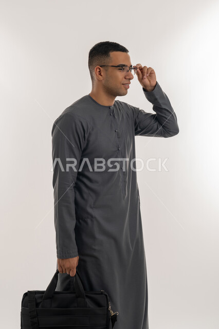 الاستعداد للعام الجامعي الجديد, التعليم العالي في الجامعات والكليات الأكاديمية الإماراتية, التحضير للذهاب إلى الجامعة, بورتريه لطالب عربي خليجي إماراتي يرتدي الكندورة يضع نظارة طبية ويحمل بيده حقيبة, الجسم بالطول الكامل, خلفية رمادية