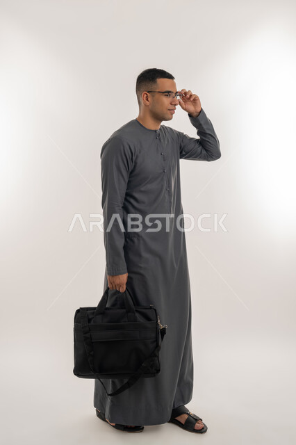 التعليم العالي في الجامعات والكليات الأكاديمية الإماراتية, التحضير للذهاب إلى الجامعة, الاستعداد للعام الجامعي الجديد, بورتريه لطالب عربي خليجي إماراتي يرتدي الكندورة يضع نظارة طبية ويحمل بيده حقيبة, الجسم بالطول الكامل, خلفية رمادية