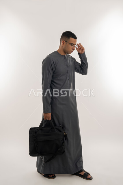التحضير للذهاب إلى الجامعة, الاستعداد للعام الجامعي الجديد, التعليم العالي في الجامعات والكليات الأكاديمية الإماراتية, بورتريه لطالب عربي خليجي إماراتي يرتدي الكندورة يضع نظارة طبية ويحمل بيده حقيبة, الجسم بالطول الكامل, خلفية رمادية