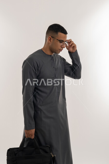 الاستعداد للعام الجامعي الجديد, التعليم العالي في الجامعات والكليات الأكاديمية الإماراتية, التحضير للذهاب إلى الجامعة, بورتريه لطالب عربي خليجي إماراتي يرتدي الكندورة يضع نظارة طبية ويحمل بيده حقيبة, الجسم بالطول الكامل, خلفية رمادية