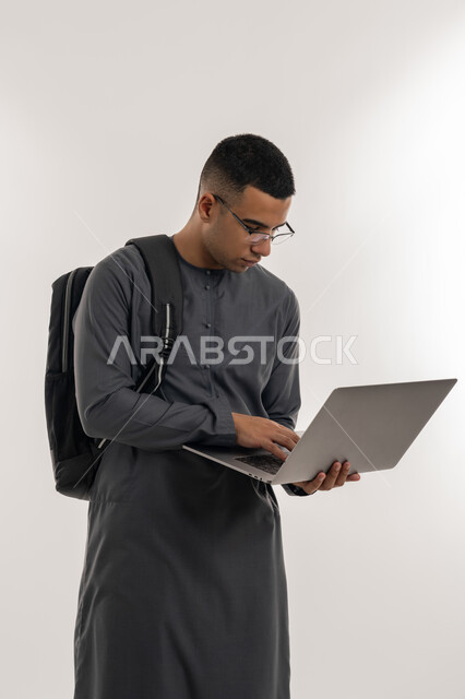 مفهوم الاجتهاد والتفوق, دمج التقنيات الحديثة بالتعليم الأكاديمي, المناهج والمواد التعليمية, بورتريه لطالب جامعي عربي خليجي إماراتي يرتدي الكندورة يحمل حقيبة على ظهره ويقوم باستعمال جهاز الحاسوب المحمول, خلفية رمادية