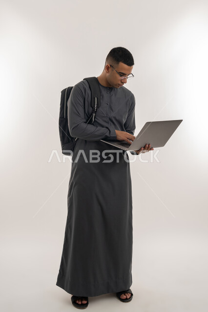 دمج التقنيات الحديثة بالتعليم الأكاديمي, مفهوم الاجتهاد والتفوق, المناهج والمواد التعليمية, بورتريه لطالب جامعي عربي خليجي إماراتي يرتدي الكندورة يحمل حقيبة على ظهره ويقوم باستعمال جهاز الحاسوب المحمول, الجسم بالطول الكامل, خلفية رمادية