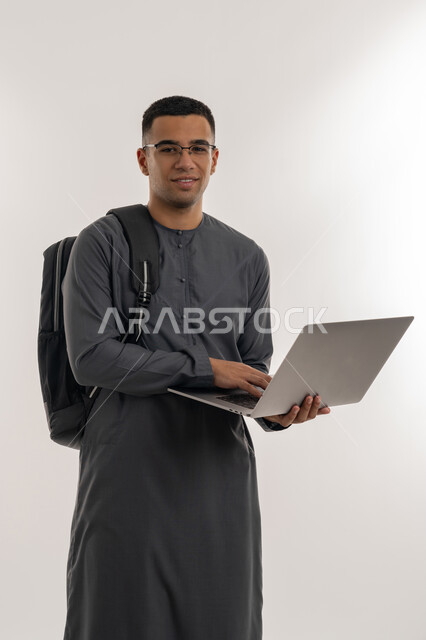 مفهوم الاجتهاد والتفوق, دمج التقنيات الحديثة بالتعليم الأكاديمي, المناهج والمواد التعليمية, بورتريه لطالب جامعي عربي خليجي إماراتي يرتدي الكندورة يحمل حقيبة على ظهره ويقوم باستعمال جهاز الحاسوب المحمول, خلفية رمادية