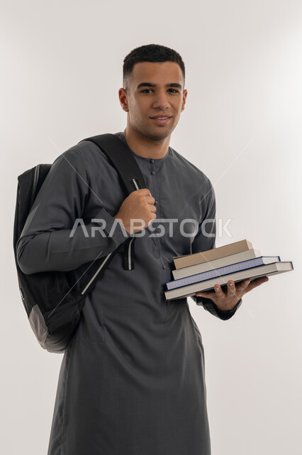 النظر إلى الكاميرا بإيماءات السعادة والسرور, التعليم العالي في الجامعات في الإمارات العربية المتحدة, الاستعداد للعام الجامعي الجديد, بورتريه لطالب جامعي عربي خليجي إماراتي يرتدي الكندورة يحمل حقيبة على ظهره وبيده الكتب, خلفية رمادية