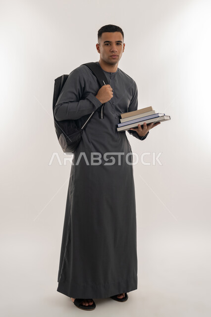 التعليم العالي في الجامعات في الإمارات العربية المتحدة, المناهج والمواد التعليمية, الاستعداد للعام الجامعي الجديد, بورتريه لطالب جامعي عربي خليجي إماراتي يرتدي الكندورة ينظر إلى الكاميرا ويحمل حقيبة على ظهره وبيده الكتب, الجسم بالطول الكامل, خلفية رمادية