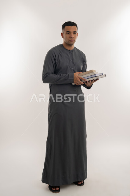 المناهج والمواد التعليمية, الاستعداد للعام الجامعي الجديد, التعليم العالي في الجامعات في الإمارات العربية المتحدة, بورتريه لطالب جامعي عربي خليجي إماراتي يرتدي الكندورة ينظر إلى الكاميرا ويحمل بيده الكتب الجامعية, الجسم بالطول الكامل, خلفية رمادية