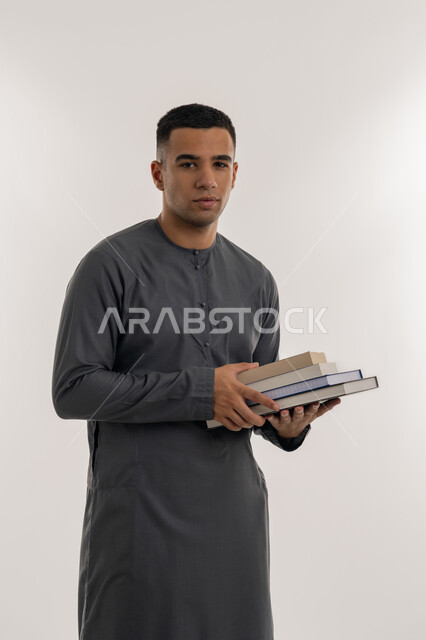 الاستعداد للعام الجامعي الجديد, التعليم العالي في الجامعات والكليات الأكاديمية في الإمارات العربية المتحدة, المناهج والمواد التعليمية الإماراتية, بورتريه لطالب جامعي عربي خليجي إماراتي يرتدي الكندورة ينظر إلى الكاميرا ويحمل بيده الكتب الجامعية, خلفية رمادية