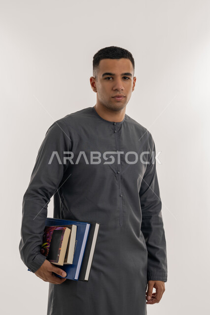 الاستعداد للعام الجامعي الجديد, التعليم العالي في الجامعات والكليات الأكاديمية في الإمارات العربية المتحدة, المناهج والمواد التعليمية الإماراتية, بورتريه لطالب جامعي عربي خليجي إماراتي يرتدي الكندورة ينظر إلى الكاميرا ويحمل بيده الكتب الجامعية, خلفية رمادية